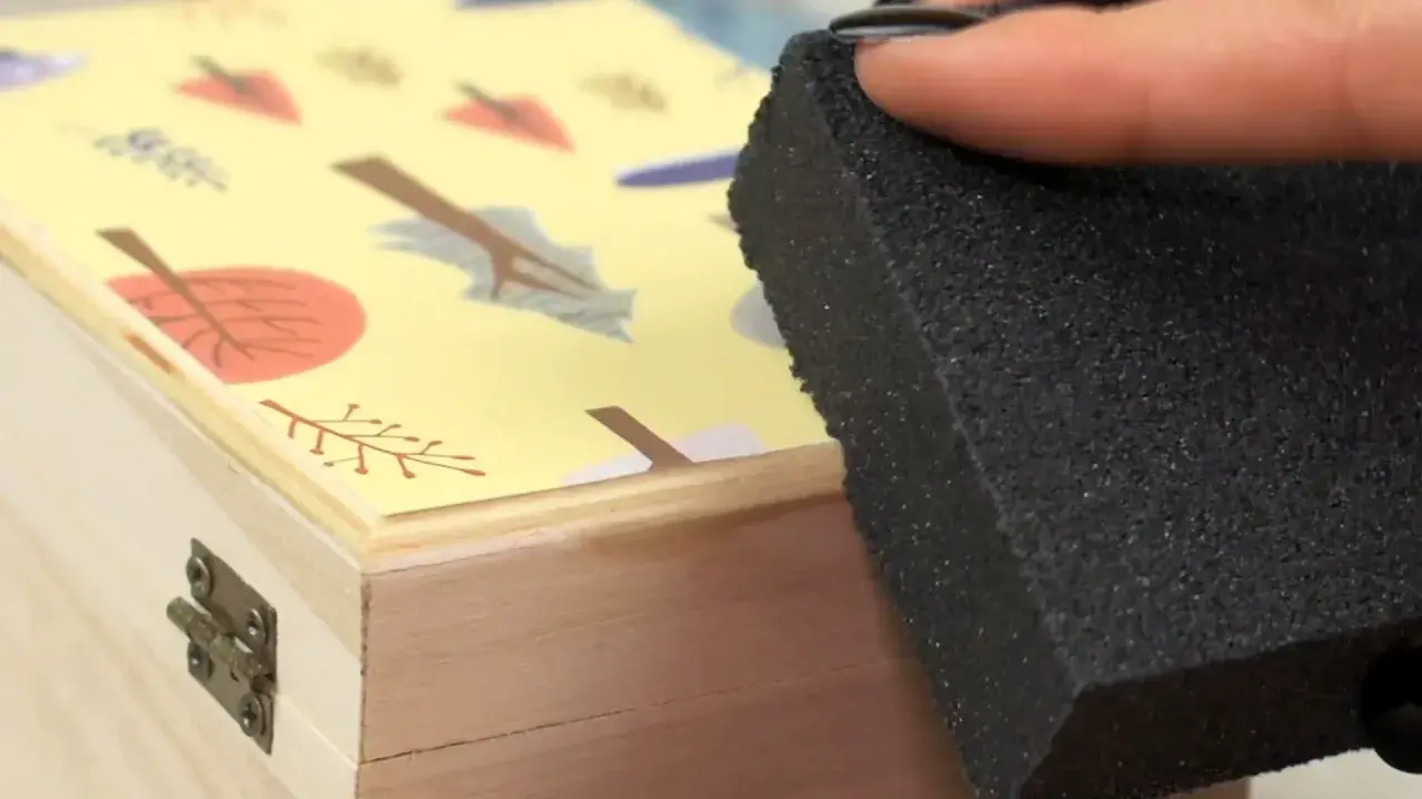 Aplicando pegamento para saber como pegar papel en madera, con una esponja sobre una caja decorada con hojas.