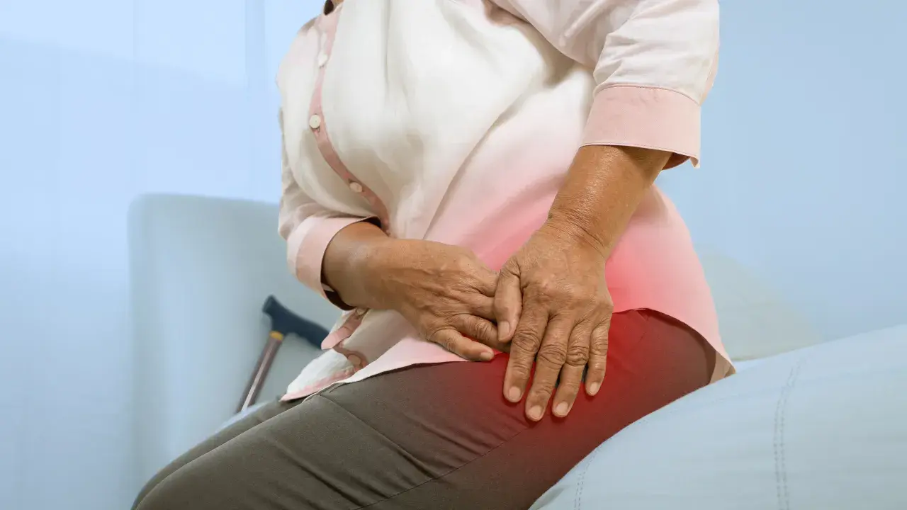 Une femme âgée se tient la hanche, souffrant de mal de hanche et sciatique. Une canne est visible derrière elle.