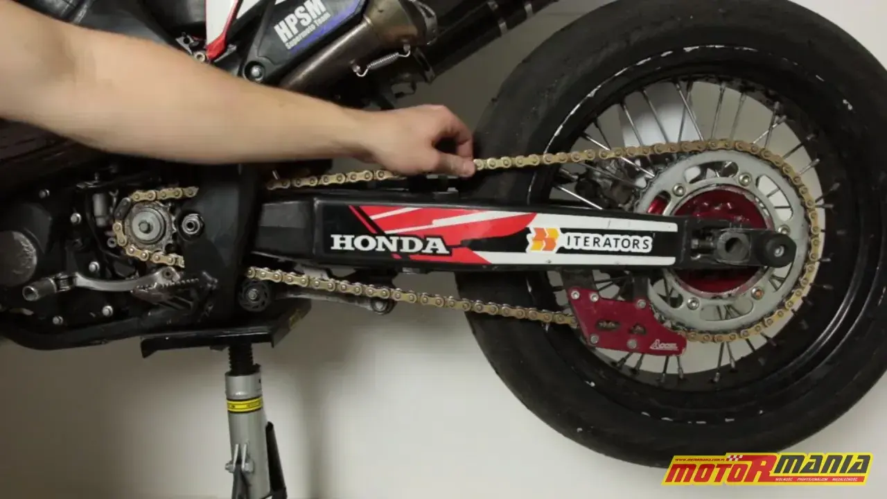 Ręka reguluje naciąg łańcucha w motocyklu Honda. Widać zębatkę, wahacz i tylne koło.