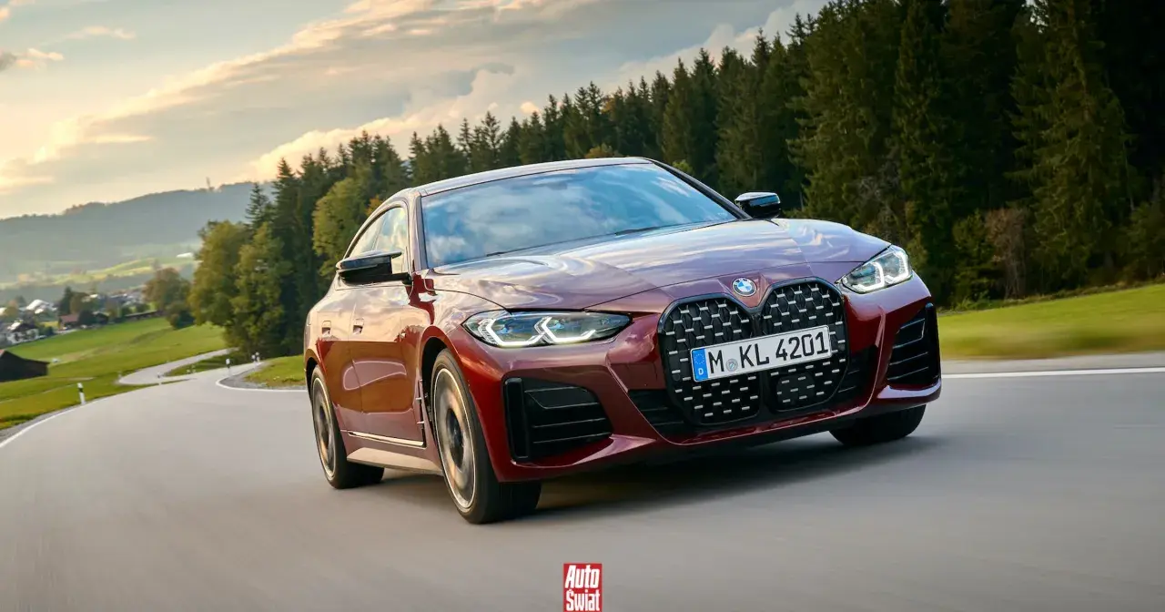 Czerwone BMW G26 z charakterystycznym grillem pędzi po krętej drodze wśród zielonych wzgórz i lasów.