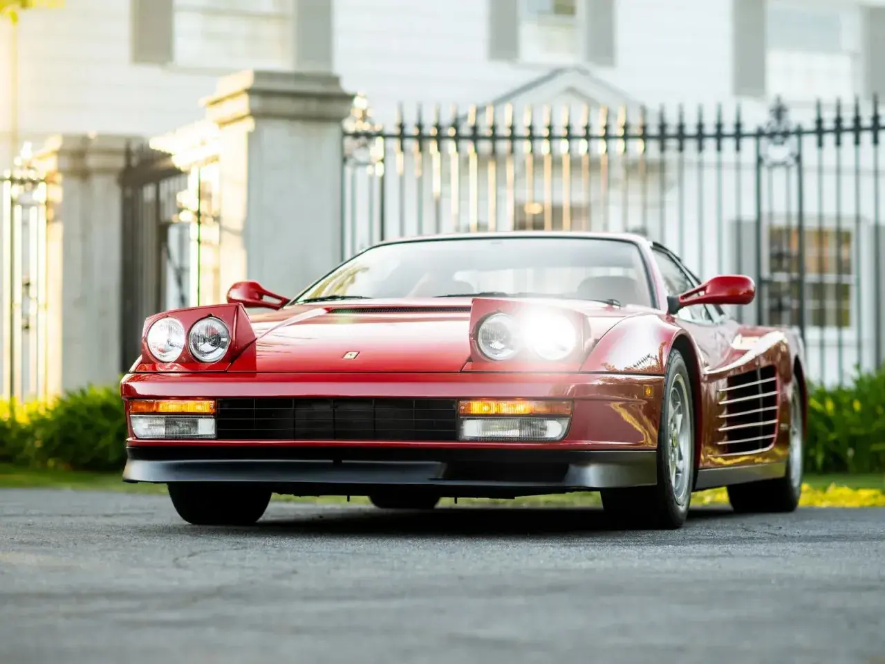 Czerwone Ferrari Testarossa z podnoszonymi światłami, zaparkowane przed elegancką posiadłością.