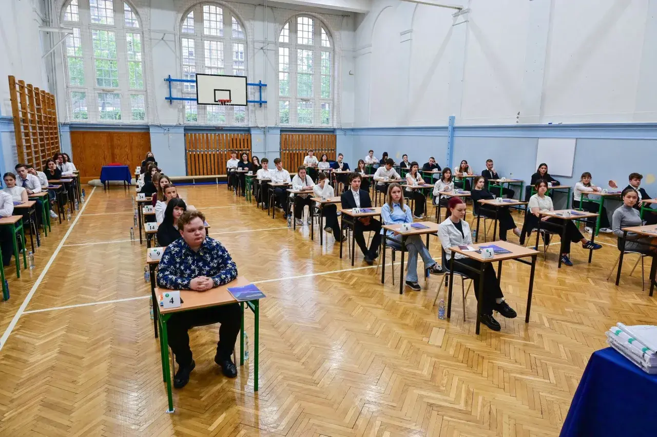 Uczniowie siedzą w ławkach w sali gimnastycznej, zdając egzamin. To typowy widok, gdy młodzi ludzie w wieku ok. 18 lat zdają maturę.