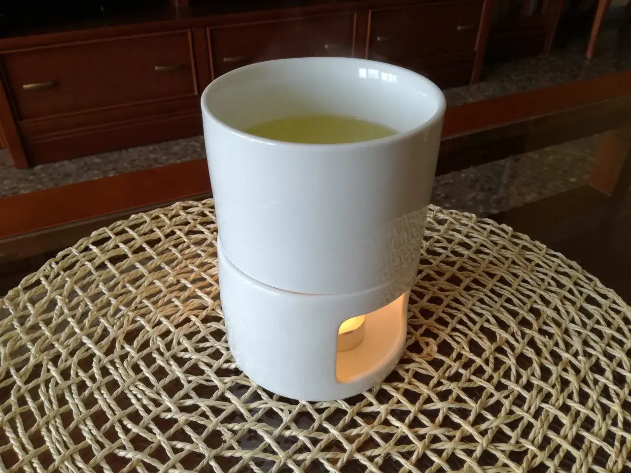 Taza blanca con líquido amarillo y vapor, sobre un soporte con vela encendida, creando un humidificador casero.