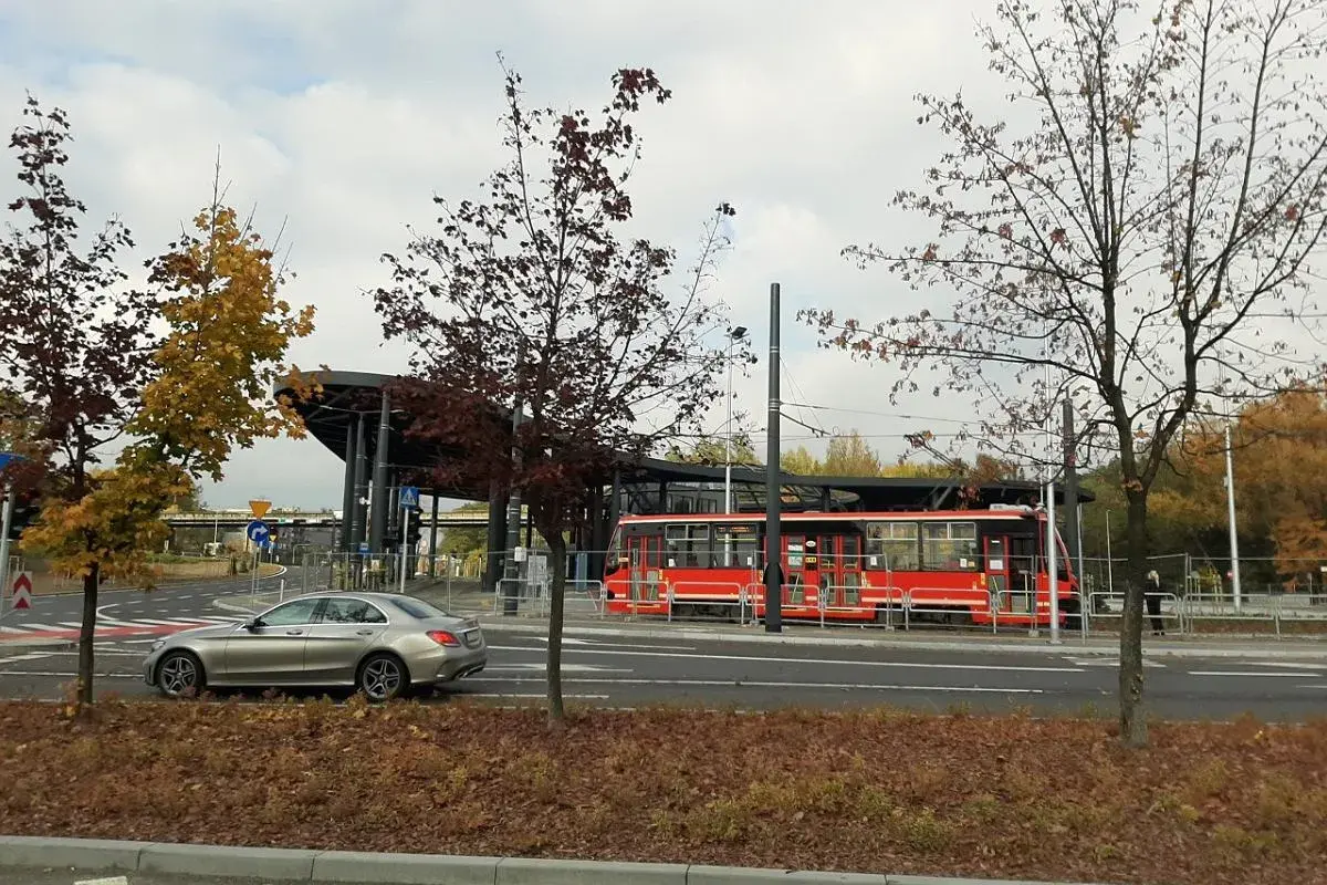 Srebrny samochód i czerwony tramwaj na przystanku, otoczone jesiennymi drzewami. Non stop big single.