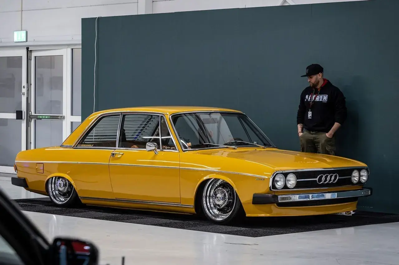 Gul Audi 100 C1, en klassiker med kromade fälgar, står parkerad bredvid en man i keps.