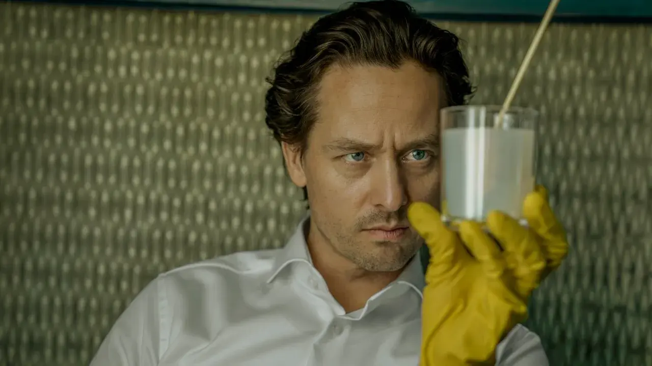 Ein Mann mit blauen Augen und gelben Handschuhen hält ein Glas mit weißer Flüssigkeit. Er ist Teil der "Achtsam Morden Reihe".