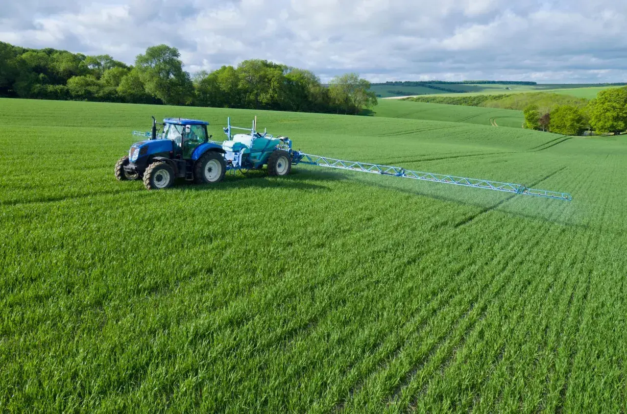 Niebieski traktor z opryskiwaczem na zielonym polu, stosuje środek Chwastox Turbo 340 SL. Dawkowanie jest kluczowe dla skuteczności.