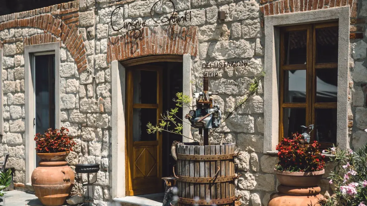 Ingresso di "Cinque Quinti", un negozio di degustazione con un torchio antico e vasi di fiori rossi.