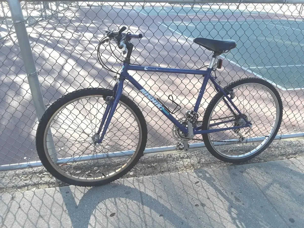 A blue Nishiki Colorado mountain bike leans against a chain-link fence, casting a shadow on the pavement.