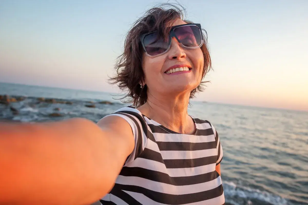 Uśmiechnięta kobieta w okularach przeciwsłonecznych robi selfie na plaży o zachodzie słońca.