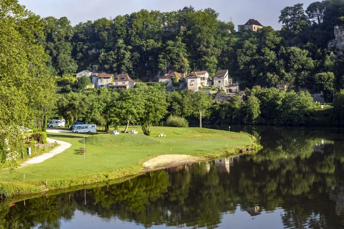 Vue paisible d'un camping-car près de la rivière, avec des maisons pittoresques accrochées à la colline, rappelant le charme de Figeac et Cahors.