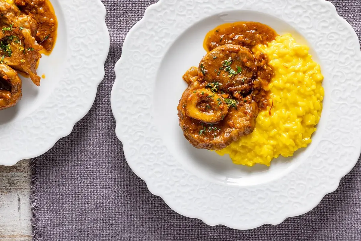 Ossobuco ricetta originale servito su un piatto bianco con risotto giallo allo zafferano.