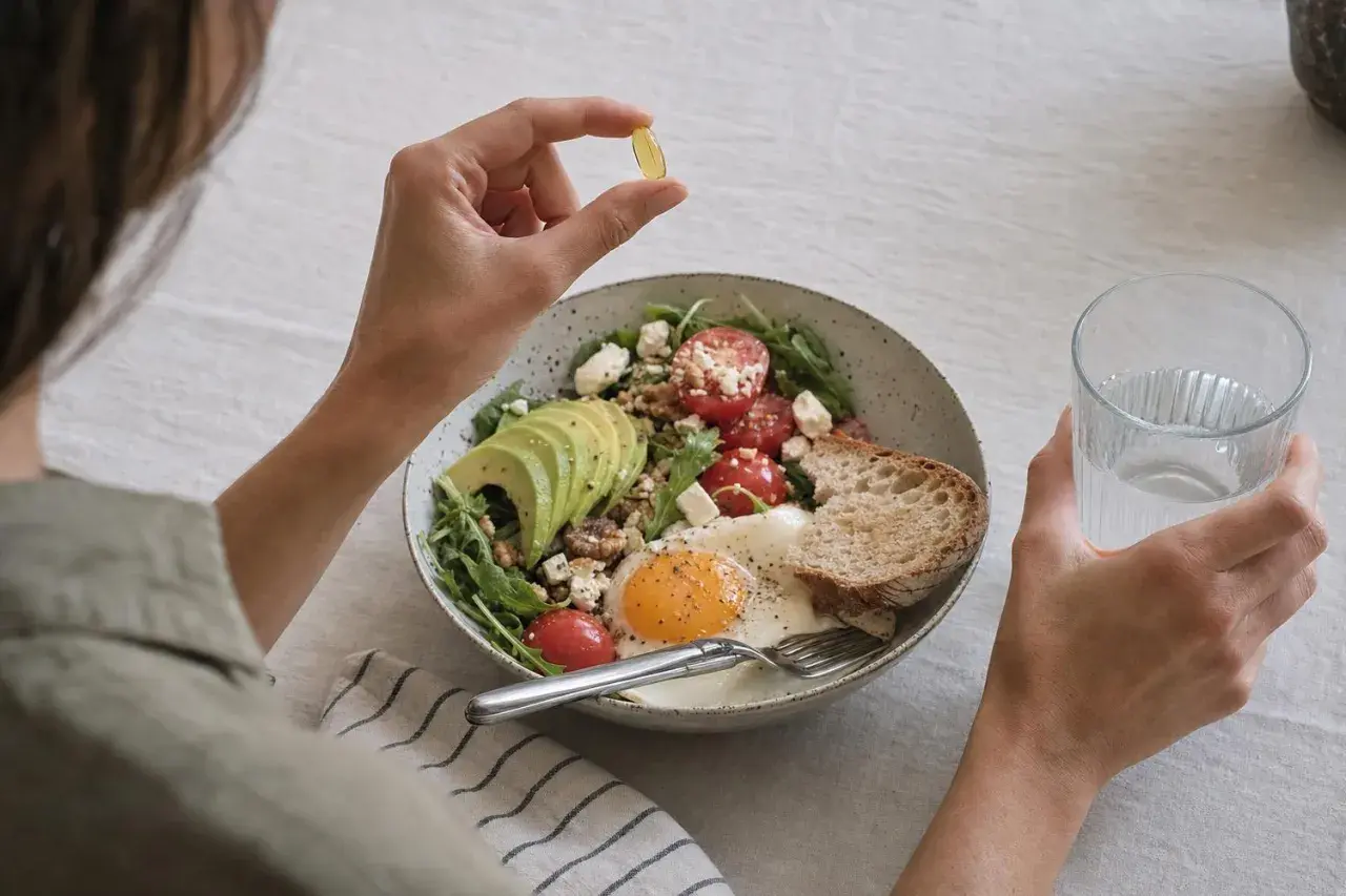 Persona prende una capsula di vitamina D, chiedendosi se sia meglio vitamina d quando prenderla mattina o sera, accanto a un piatto salutare.