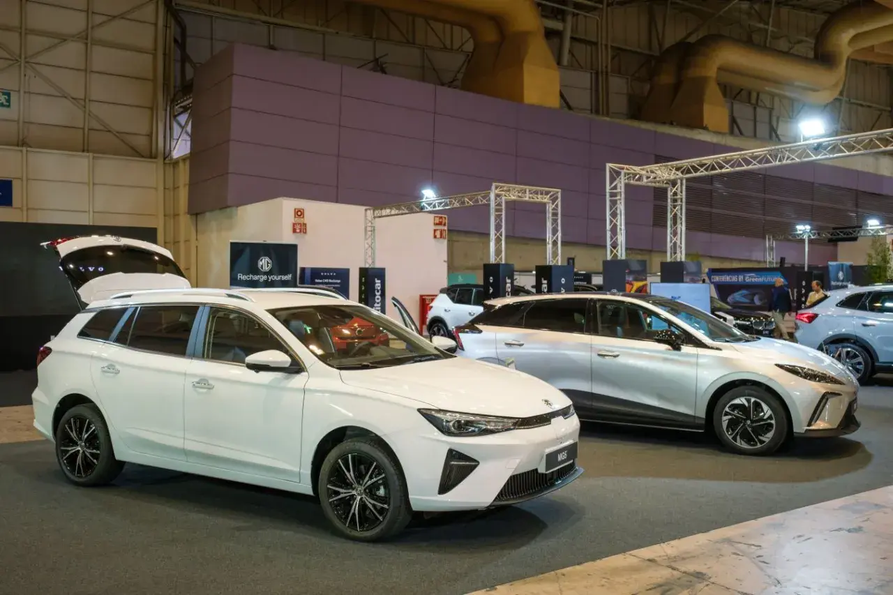Białe kombi MG5 z otwartą klapą bagażnika i srebrny SUV MG na wystawie samochodowej.
