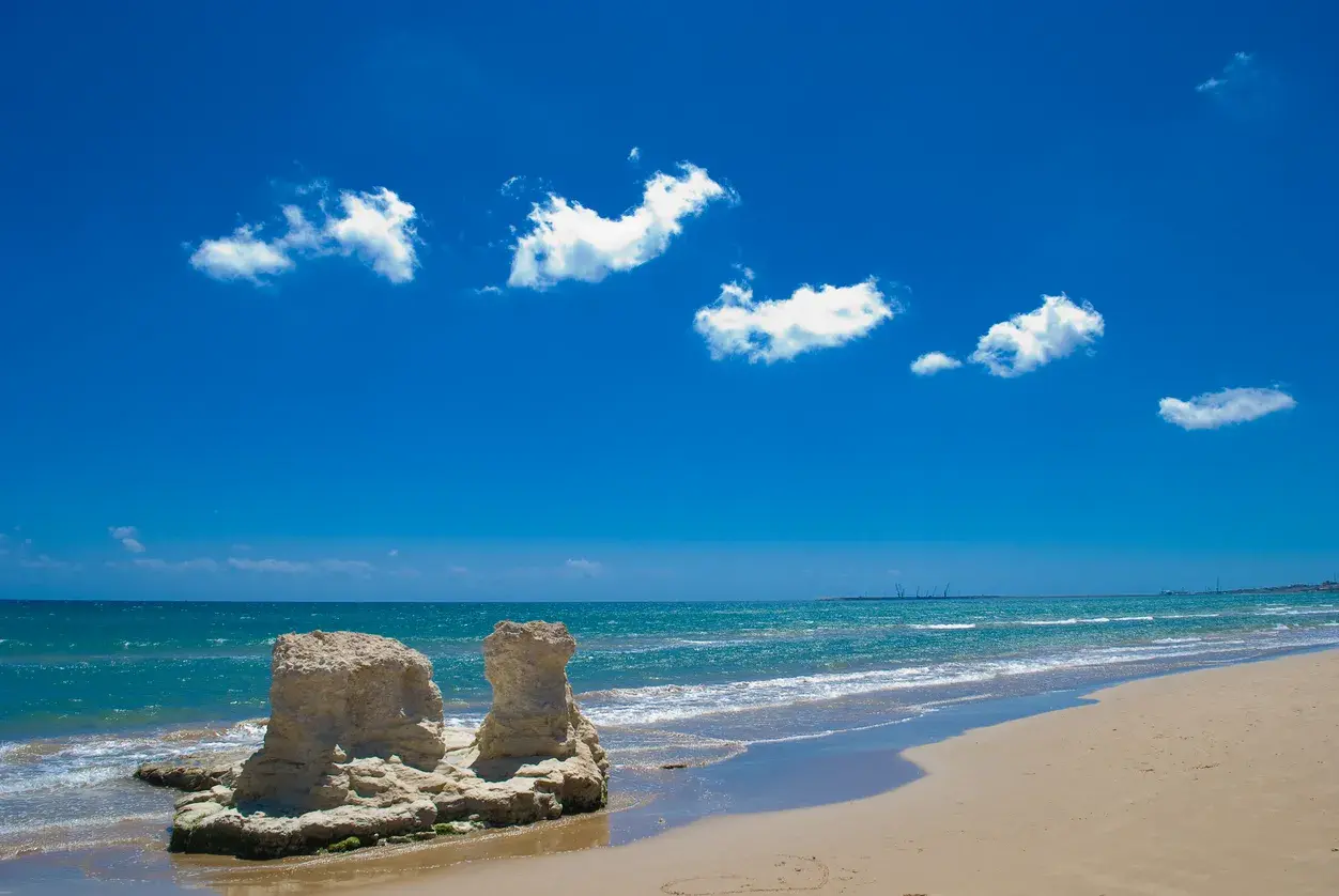 Spiagge libere Sicilia: rocce scolpite dal mare e sabbia dorata sotto un cielo azzurro con nuvole bianche.