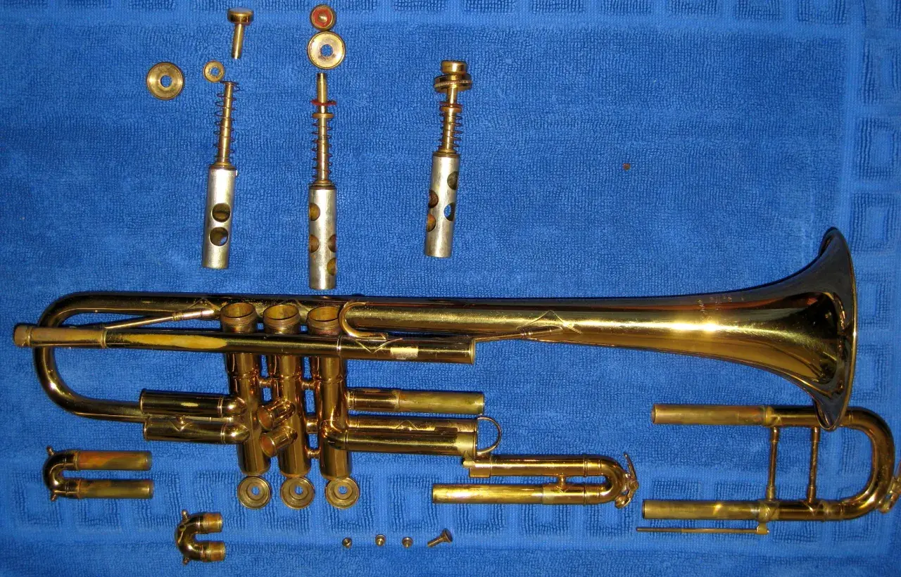 Rozłożona trąbka, instrument dęty blaszany, leży na niebieskim tle. Widać jej poszczególne części: korpus, wentyle, ustnik i inne elementy.