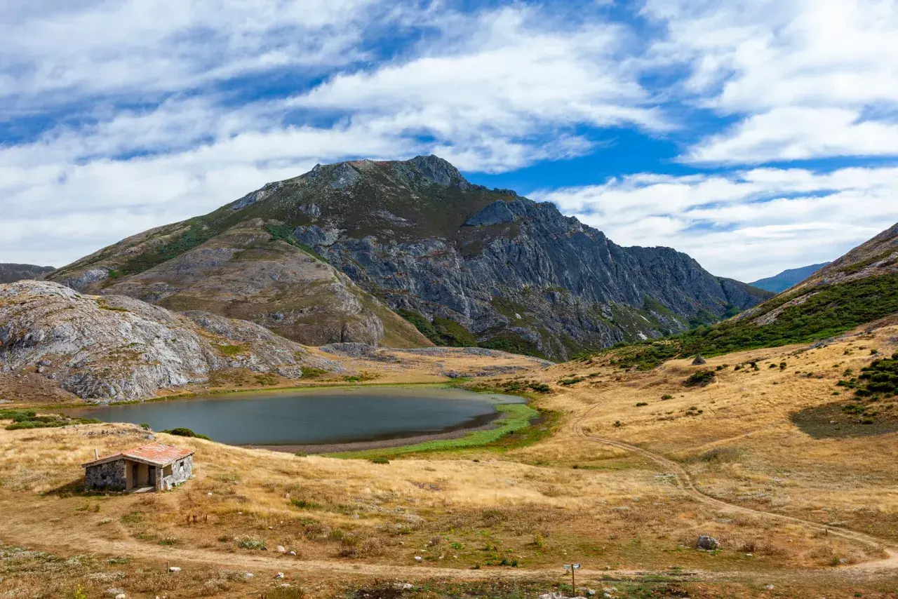 Pequeña cabaña de piedra junto a un lago ausente, rodeada de montañas escarpadas y prados secos bajo un cielo azul con nubes.