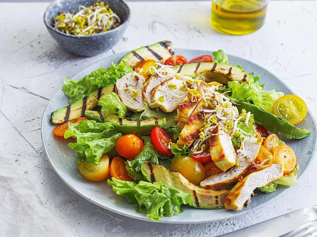 Gegrillter Hähnchensalat mit Avocado, Tomaten und Sprossen auf einem Teller.