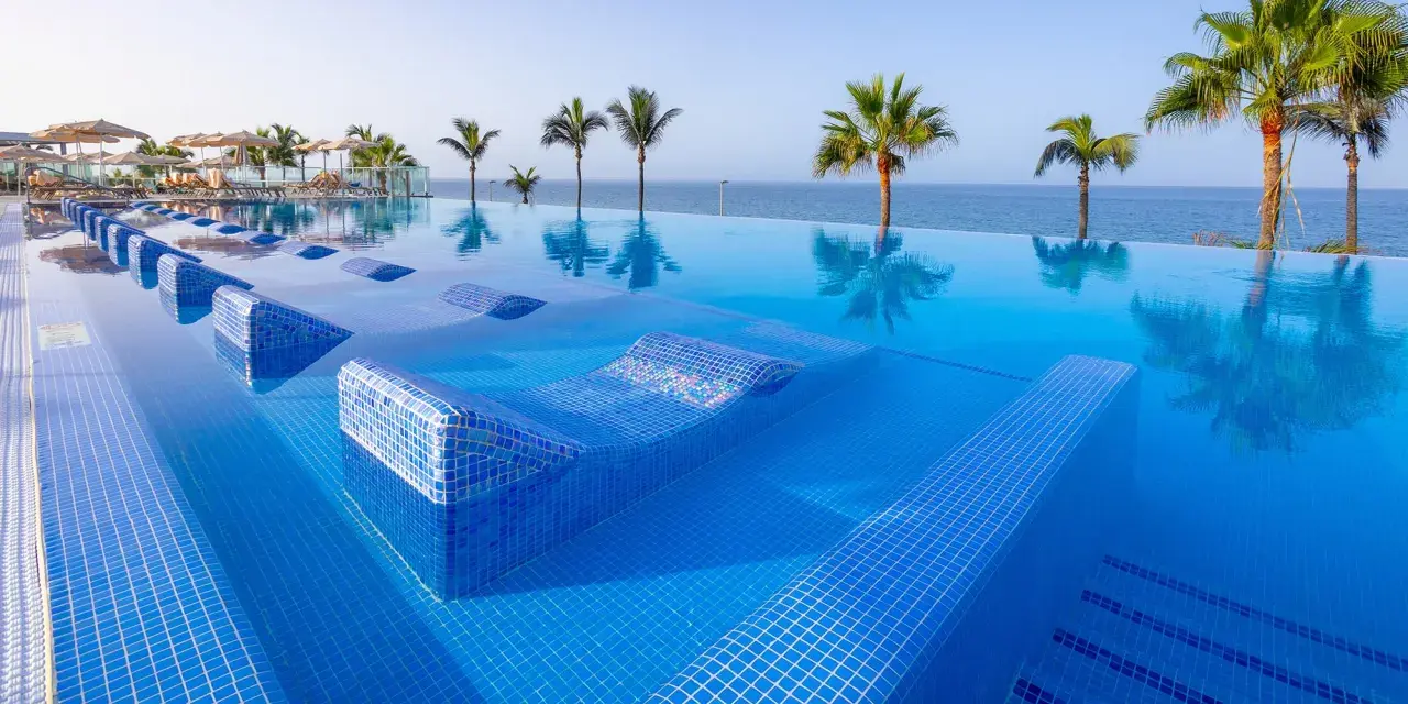 Infinity-Pool mit integrierten Liegen im Riu Gran Canaria, Palmen und Meer im Hintergrund.