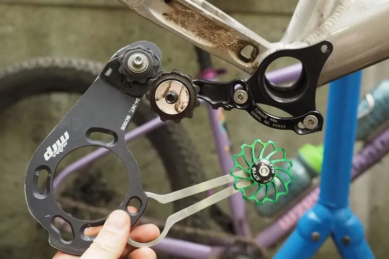 A hand holds a bike chain guide, part of a 1x drivetrain, near a bicycle frame. A green pulley wheel is also visible.