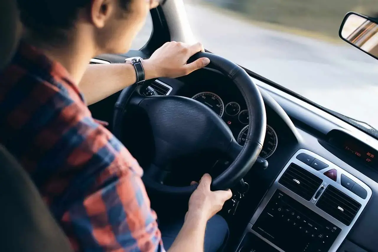 Uomo al volante di un'auto, con le mani sul volante. La sua camicia a quadri rossi e blu si abbina al cruscotto dell'eps auto.