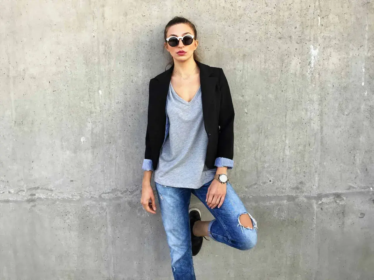 Stylisches Outfit mit Blazer: Frau in Jeans, grauem V-Shirt und Sonnenbrille lehnt an Betonwand.