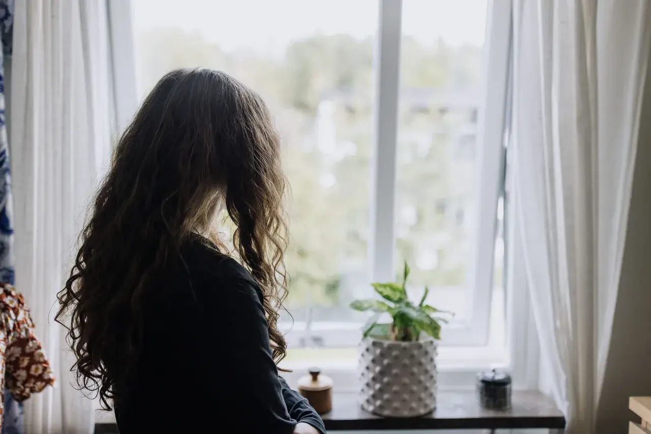 Flicka med långt hår tittar ut genom fönstret. En planta står på fönsterbrädan. Bilden kan symbolisera emotionell utveckling 3 år.