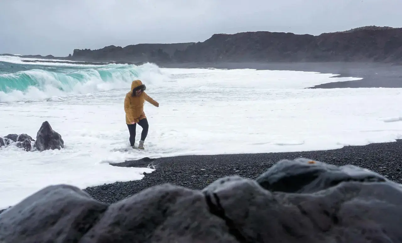 Kobieta w żółtej kurtce brodzi w pianie na czarnej plaży Reynisfjara, podczas gdy potężne fale rozbijają się o brzeg.