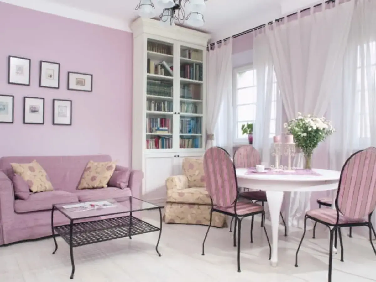 Jasny salon z fioletową kanapą i krzesłami w różowe pasy. Kolor lawendowy ścian świetnie komponuje się z bielą mebli i zasłon, tworząc przytulną atmosferę.