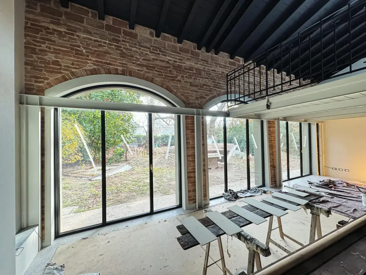 Interno in fase di ristrutturazione con ampie vetrate sul giardino. In primo piano, assi di legno appoggiate su cavalletti, segno di lavori di miglioramento della casa in corso.