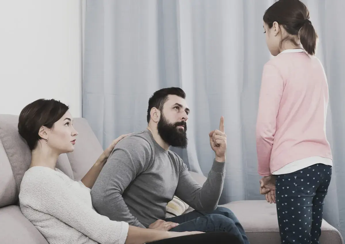 Una bambina ascolta il padre che la ammonisce, mentre la madre, con un'espressione preoccupata, le appoggia una mano sulla spalla, forse per alleviare la sua paura di perdere i genitori.