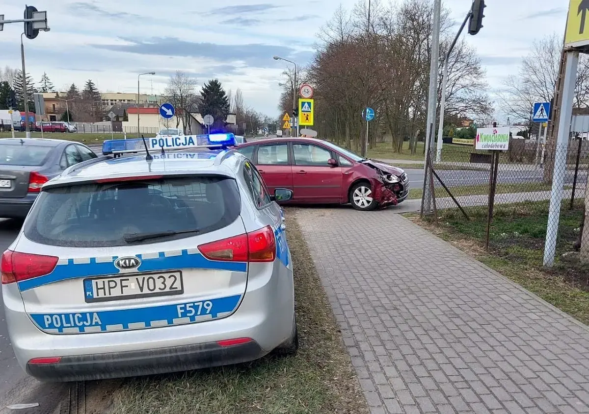 Samochód policyjny z włączonymi sygnałami świetlnymi przy drodze, gdzie doszło do kolizji drogowej. Czerwony samochód ma uszkodzony przód.