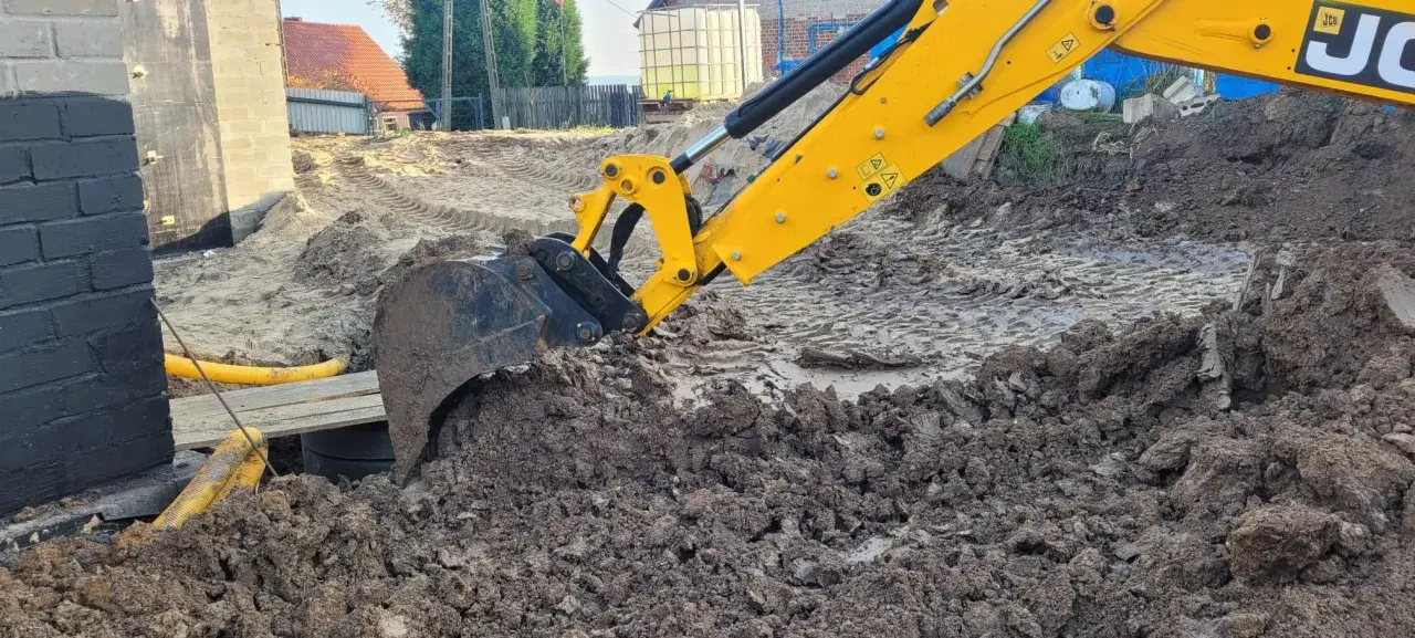 Żółta koparka marki JCB pracuje na budowie, kopiąc ziemię. Widać jej ramię i łyżkę zagłębiającą się w błotniste podłoże.