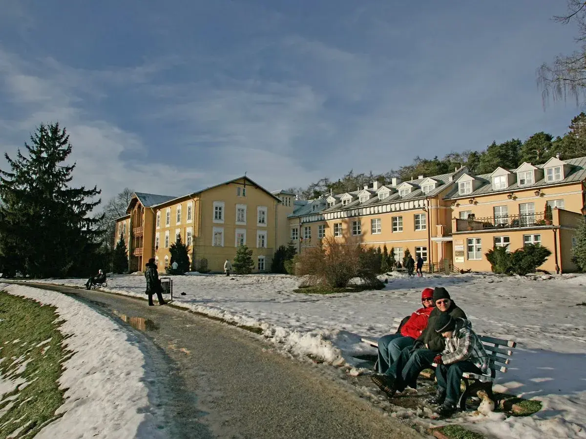 Żółty budynek sanatorium otoczony śniegiem, ludzie siedzą na ławce.