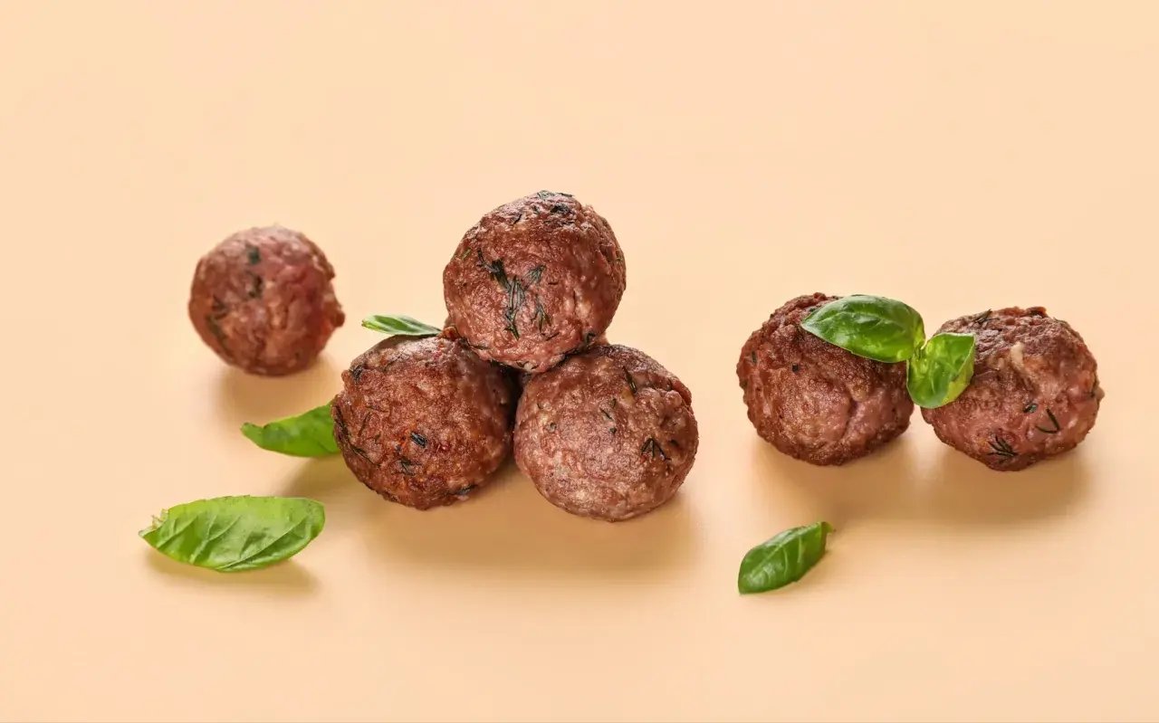 Cinq boulettes de viande dorées, garnies de feuilles de basilic frais, sur fond beige.