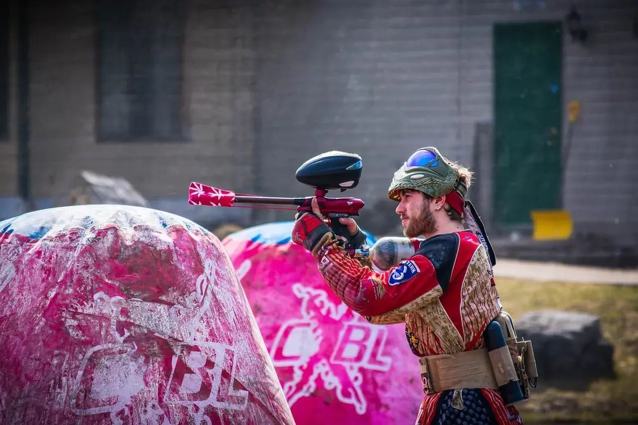 Gracz w stroju bojowym celuje z markerem, gotowy do akcji w laserowym paintballu. Za nim różowe dmuchane przeszkody.