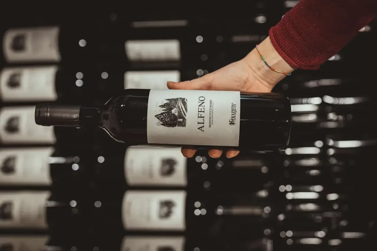 Mano che tiene una bottiglia di vino rosso, etichetta "Alferno Montecucco Rosso", un vino che ricorda il cabernet.