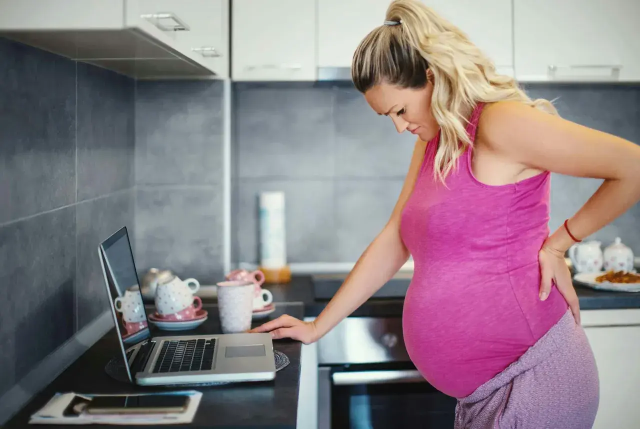 Gravid kvinna med ont i ryggen, hon kan inte gå p.g.a. foglossning. Hon står vid en bänk med en laptop.