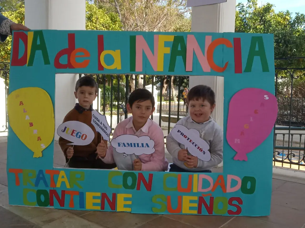 Niños sonríen tras un marco decorado para el Día de la Infancia, sosteniendo carteles con palabras como "Juego", "Familia" y "Protección".