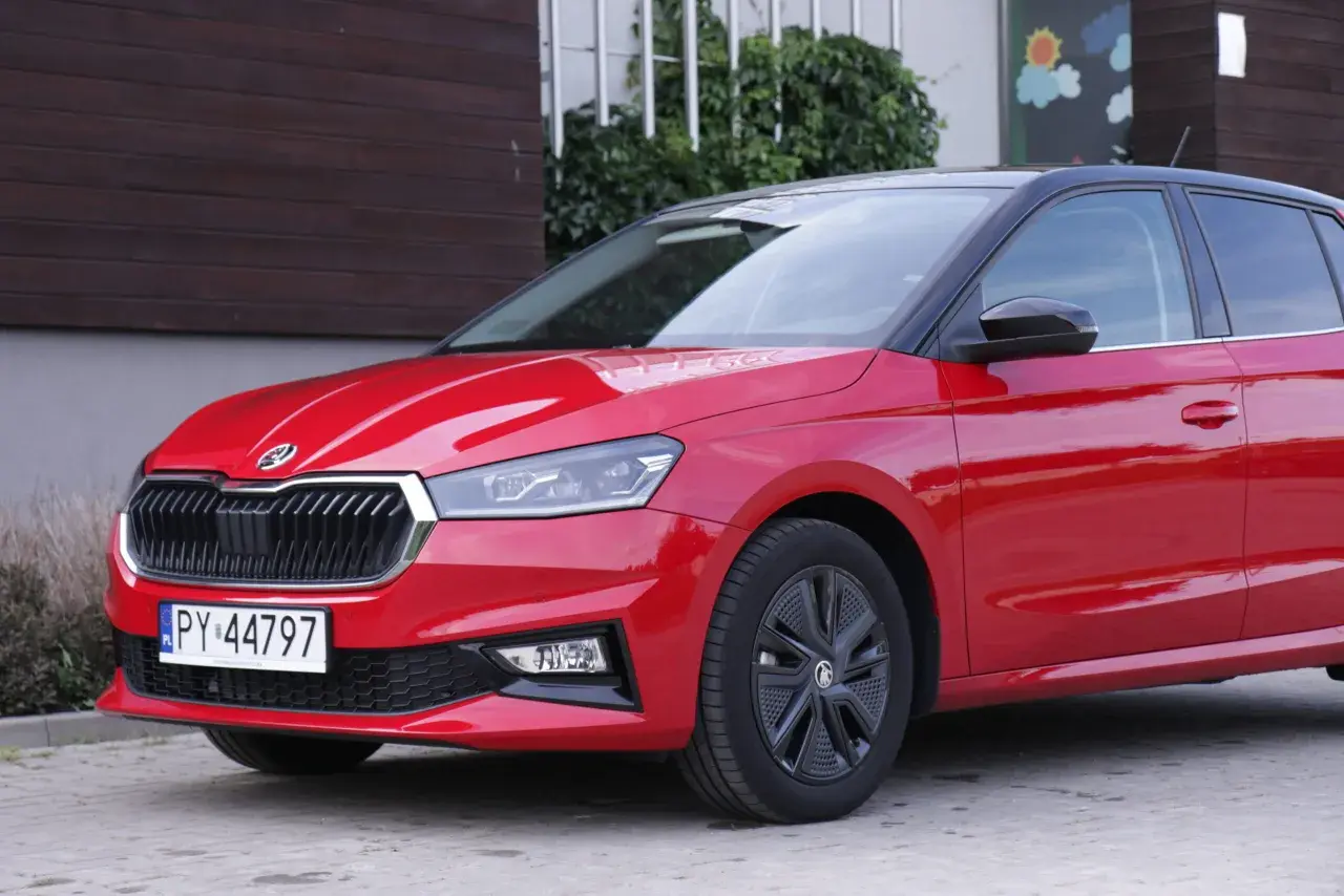 Czerwona Skoda Fabia 4 z czarnym dachem i felgami stoi przed budynkiem z drewnianą elewacją.