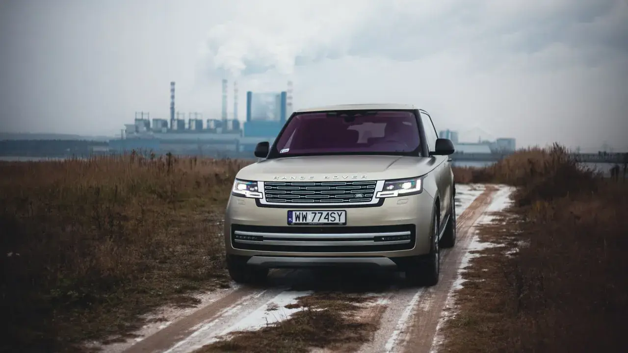 Złoty Range Rover Autobiography na błotnistej drodze, w tle przemysłowy krajobraz z kominami fabrycznymi.