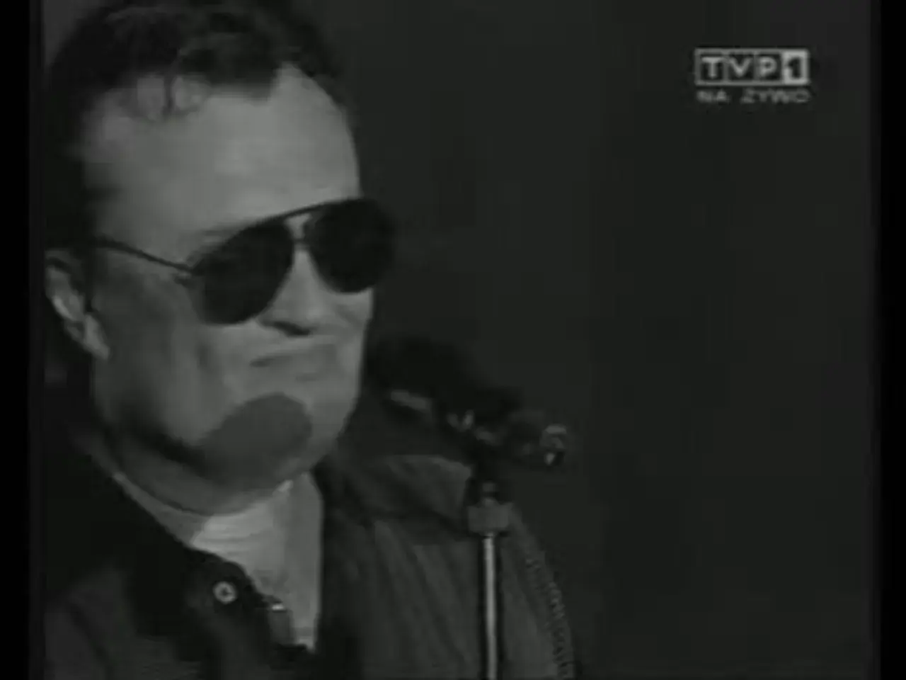 Mężczyzna w okularach przeciwsłonecznych śpiewa do mikrofonu. Widać logo TVP1. A po nocy przychodzi dzień.