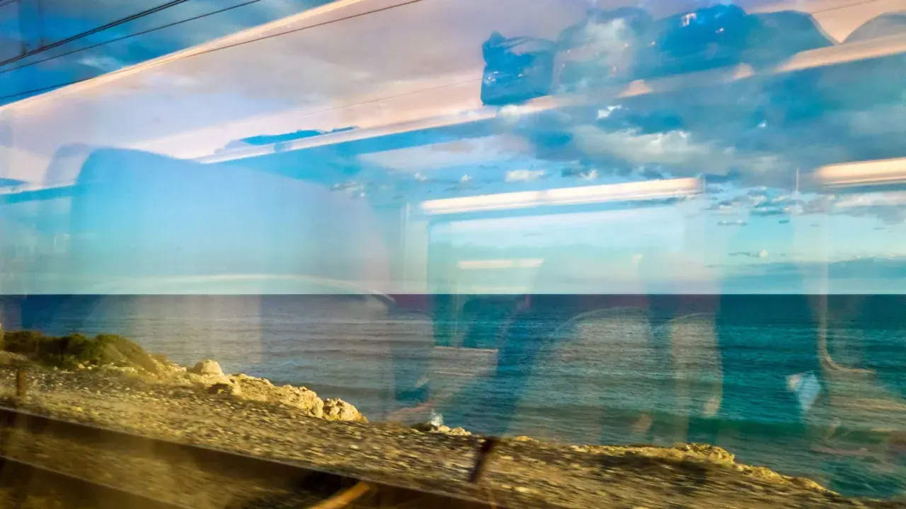Blick aus dem Zugfenster auf das Meer. Die Spiegelungen im Glas zeigen Wolken und den Himmel, während draußen die Küste vorbeizieht. Mit dem Zug ans Meer ist ein Erlebnis.