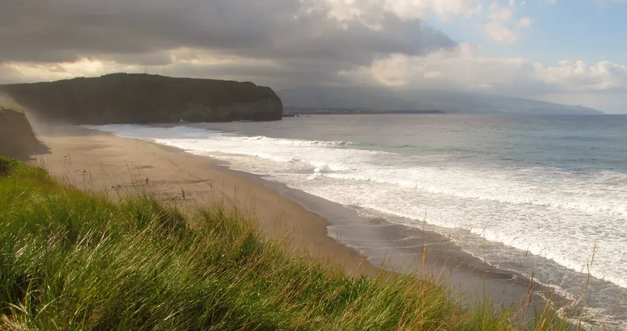 Ein einsamer azoren strand mit sanften Wellen und dramatischen Wolken. Hohes Gras wiegt sich im Wind.