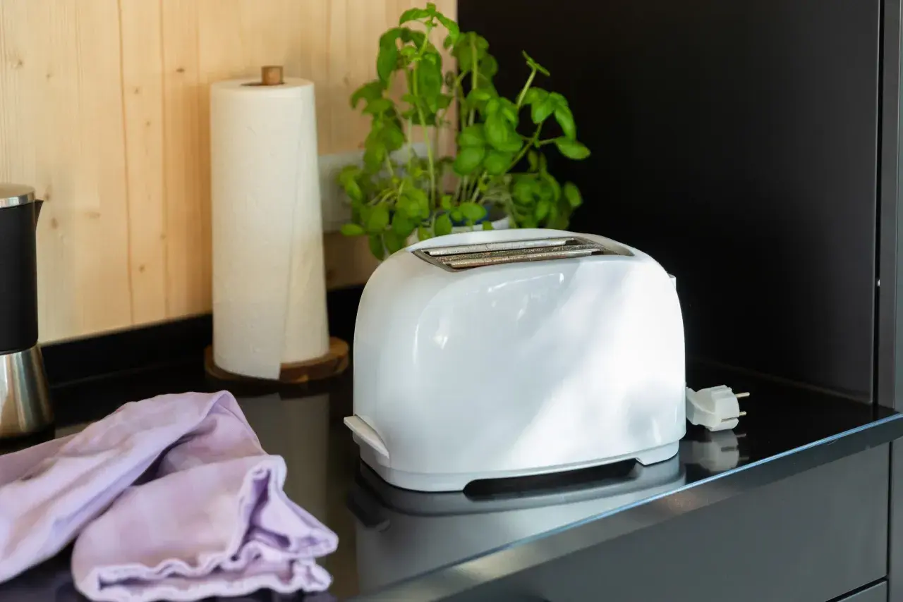 Ein weißer Toaster steht auf einer schwarzen Arbeitsplatte, bereit für die Reinigung. Daneben liegen ein lila Tuch und eine Küchenrolle.