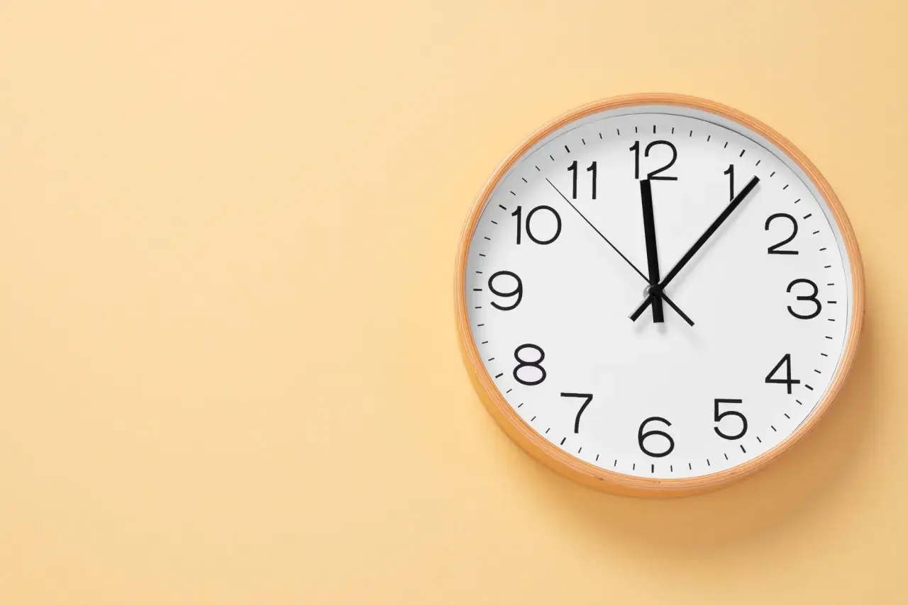 Reloj de pared blanco con marco de madera. Las manecillas marcan las 12:05. Ideal para aprender las horas en inglés, ejemplos.