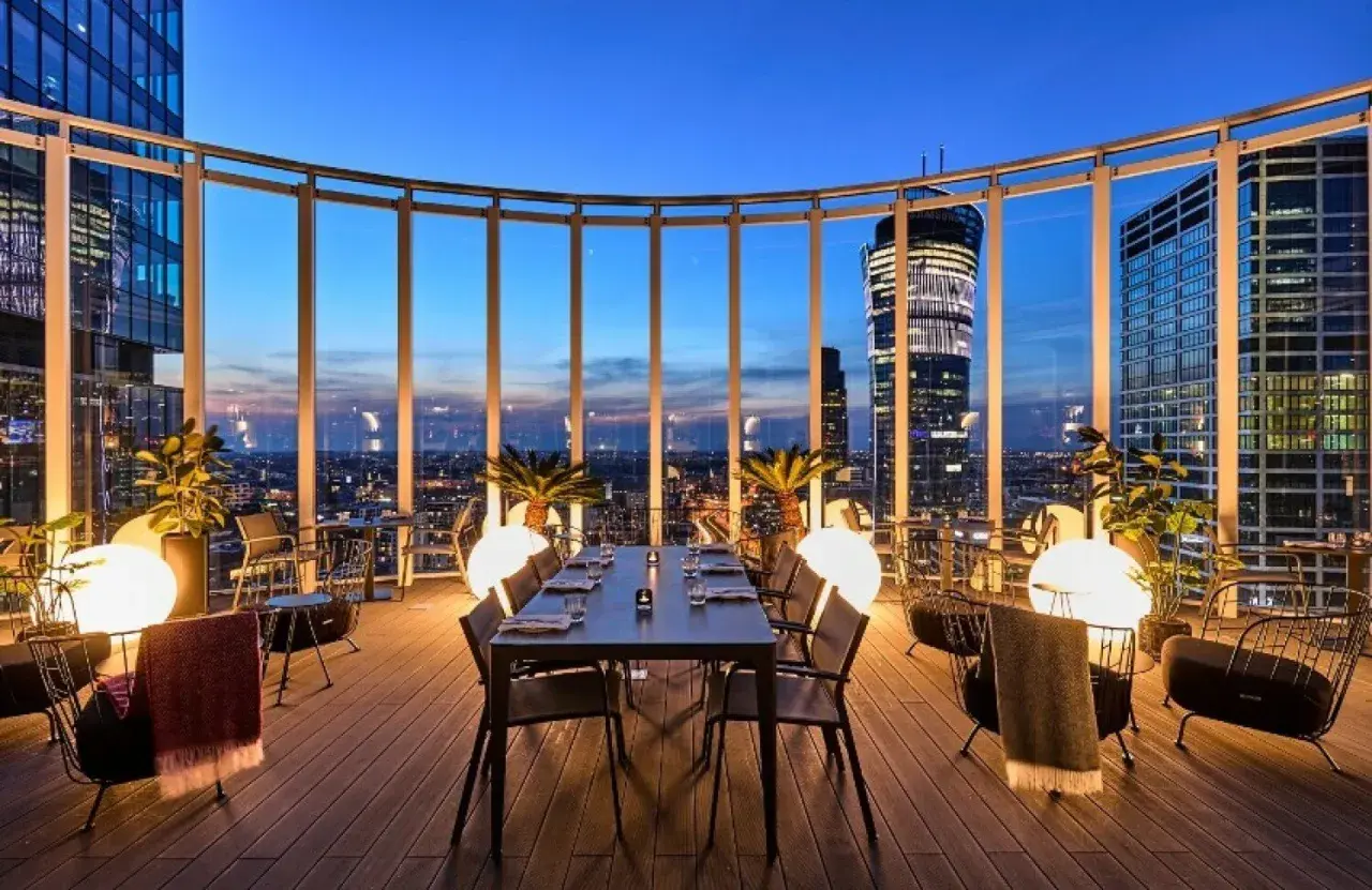 Przytulna restauracja Warszawa na dachu z widokiem na nocne miasto. Stoły i krzesła, palmy i kule świetlne tworzą magiczną atmosferę.