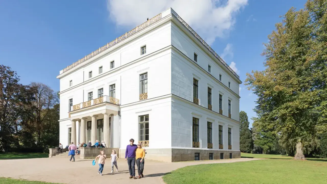 Familie spaziert durch den Jenisch Park vor einem weißen Herrenhaus mit Säulen und goldenen Verzierungen.