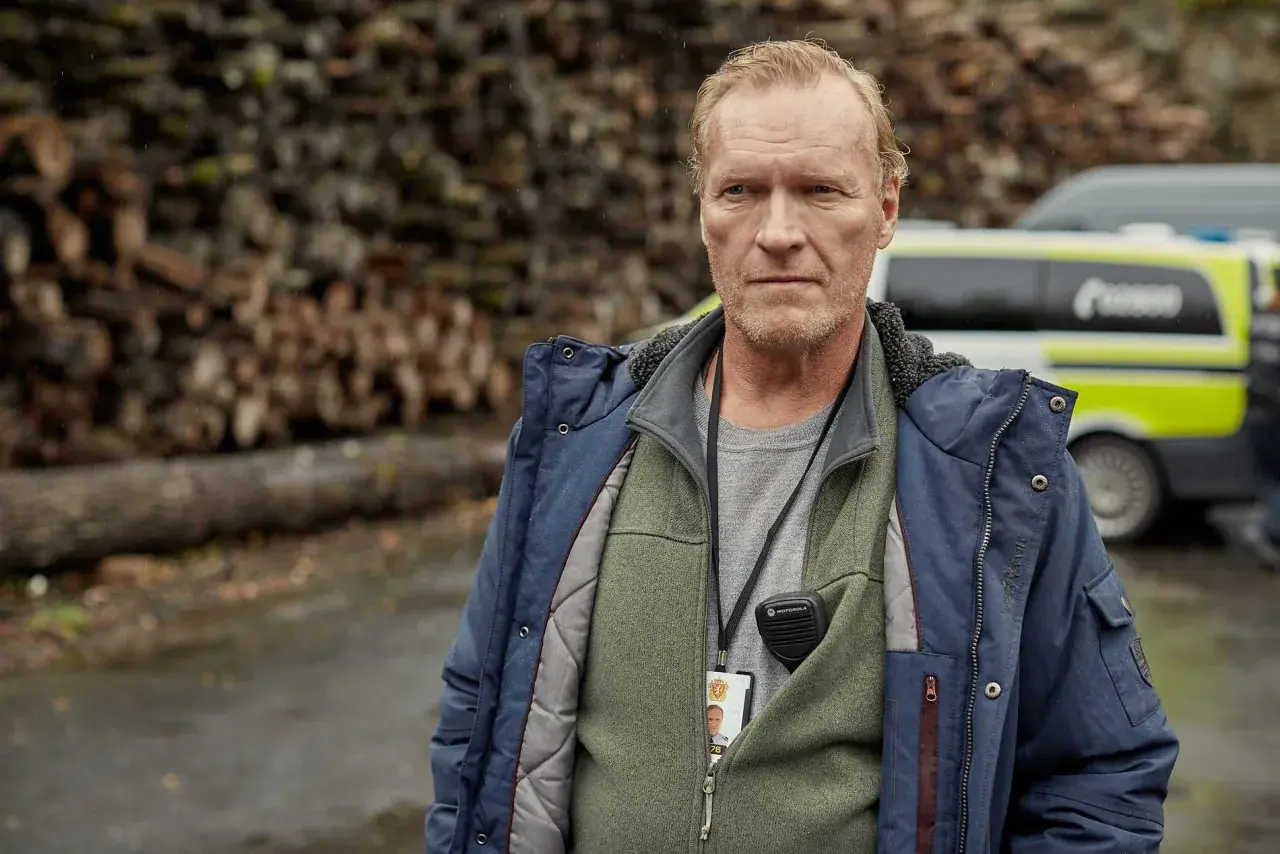 En man med intensiv blick, klädd i jacka och väst, bär en radio och ett id-kort. Bakom honom syns en polisbil och en vedhög. En scen från en norsk serie.