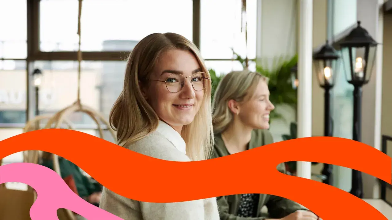 Två leende kvinnor sitter vid ett bord. Den ena tittar mot kameran. Kanske funderar de på vad som går arbetsgivaravgiften till.